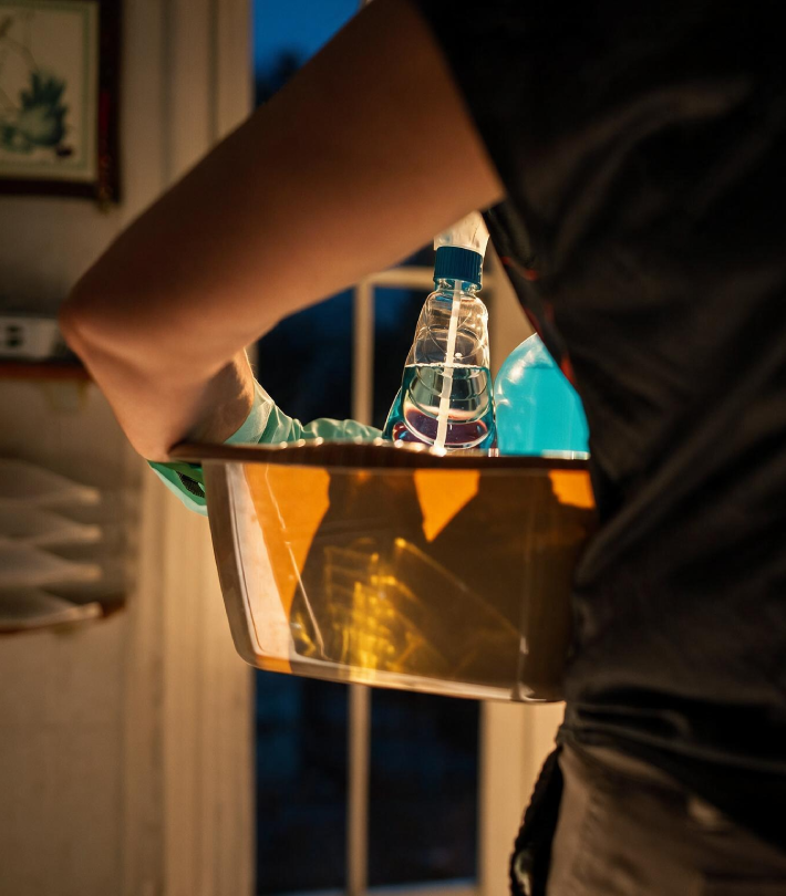 Janitor carrying a yellow caddy filled with spray bottles and cleaning products near a window at night.